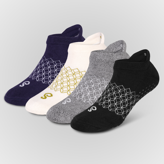 Four pairs of low-cut ankle grip socks arranged in a fan shape on a light gray background. The socks come in navy blue, white, heather grey, and black, each featuring a white geometric honeycomb pattern on the foot and a small white stylized logo near the toe.